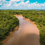 Vista aérea de uma embarcação navegando por um rio de águas barrentas, cercado pela floresta amazônica.