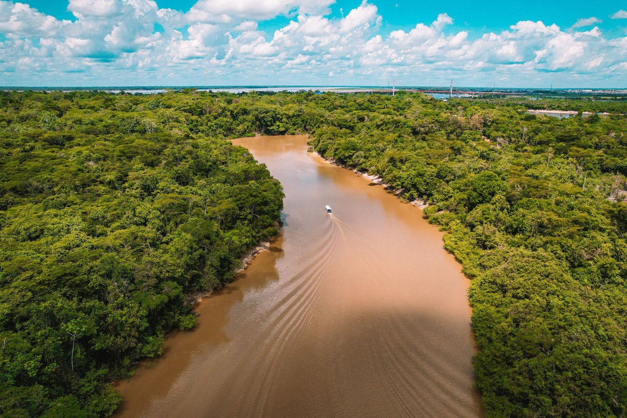Vista aérea de uma embarcação navegando por um rio de águas barrentas, cercado pela floresta amazônica.