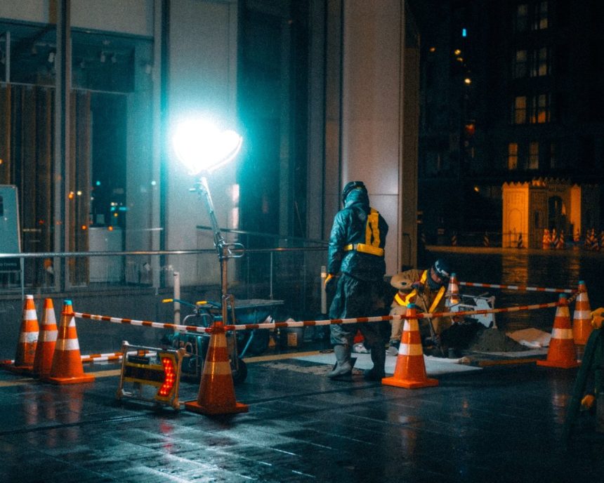 Trabalhadores da construção caminham à noite em uma área urbana iluminada.